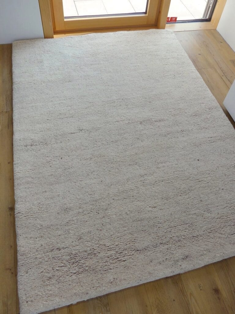 Een beige rechthoekige Houseify Handmade | Berber Vloerkleed | Gemêleerde wit wordt op een houten vloer bij een raam geplaatst.