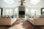 Een lichte woonkamer met twee beige banken tegenover elkaar, een tv boven een open haard, een glazen salontafel, dakramen, grote ramen, planten en een organisch vloerkleed op de vloer.