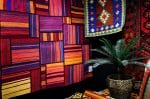Er wordt een levendige tentoonstelling van kleurrijke geweven tapijten met geometrische patronen getoond, waarbij een kleed aan de muur een centraal punt vormt. Ervoor staat een palmplant in pot, die een vleugje groen aan het tafereel toevoegt.