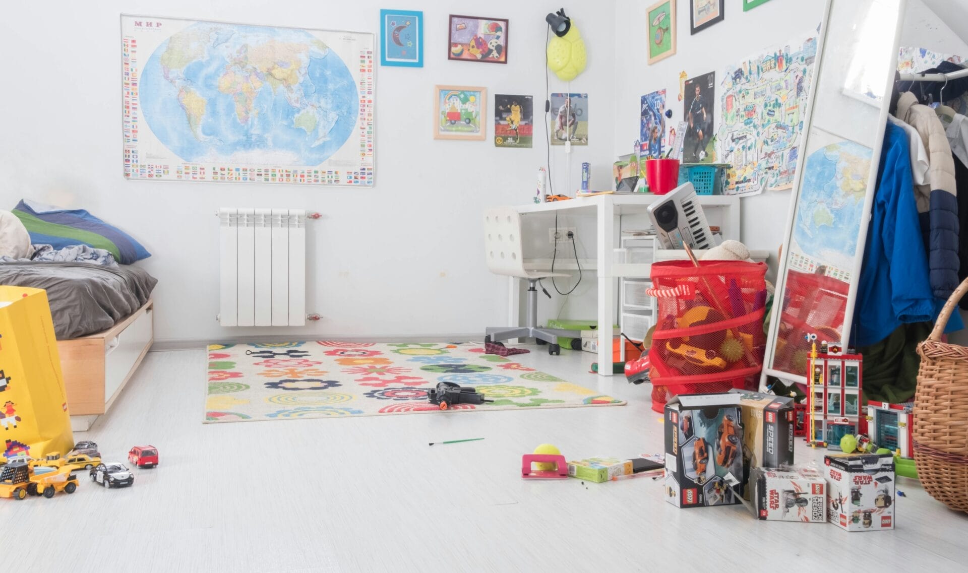 Een kinderslaapkamer met een doordachte babykamerindeling met links een bed, rechts een bureau en verspreid op de vloer divers speelgoed. Aan de muren hangen kaarten, posters en charmante tekeningen.