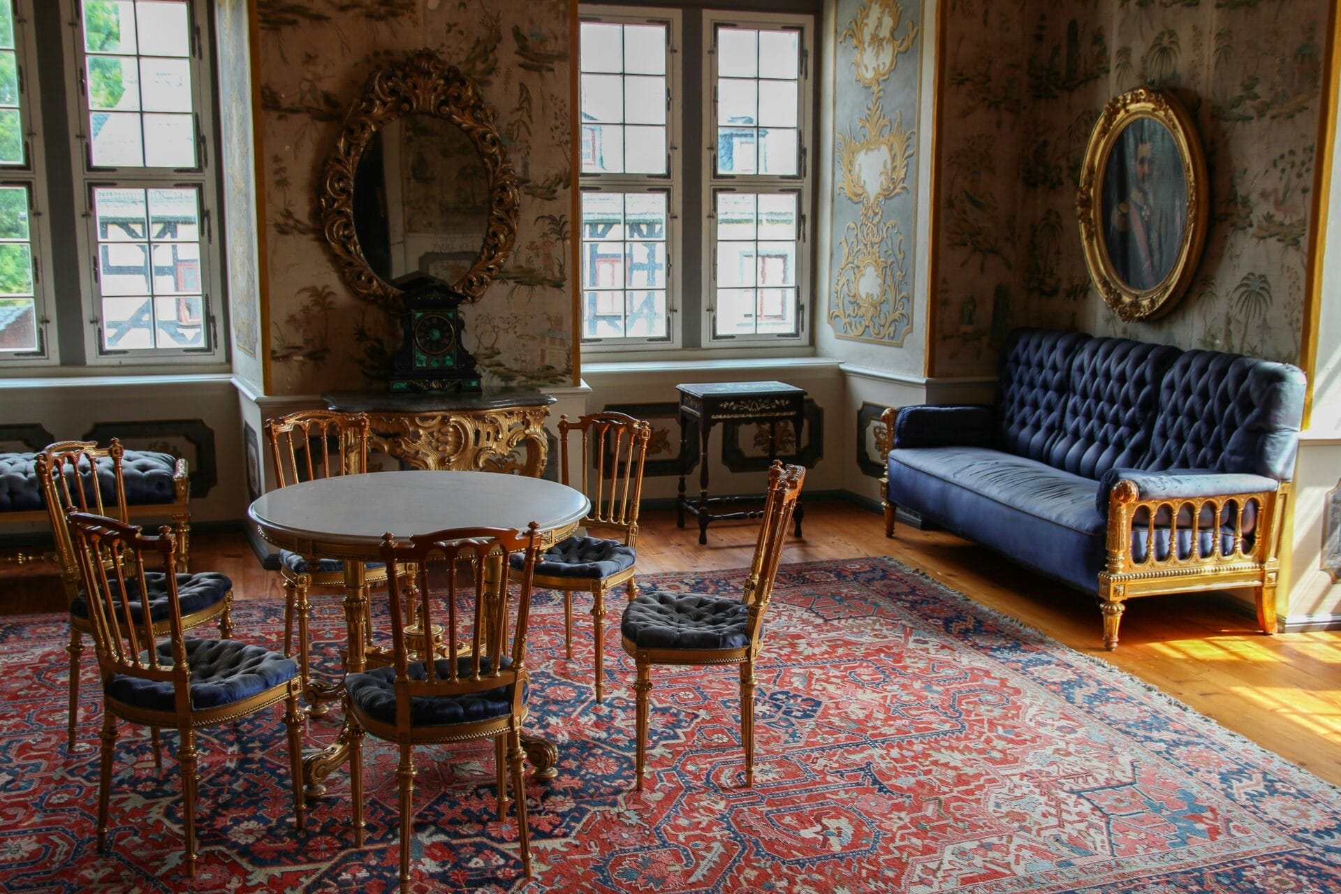 Een kamer met sierlijk meubilair, waaronder een ronde tafel met vijf stoelen, een blauwe getufte bank, een anti-allergietapijt, een gedecoreerde spiegel boven een open haard en schilderijen aan de muren.