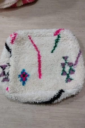 Een vierkante, donzige witte Marokkaanse Beni Ouarain Berber Poef 010 met kleurrijke abstracte patronen, waaronder vormen en lijnen in roze, blauw, groen, zwart en paars, liggend op een lichte houten vloer.