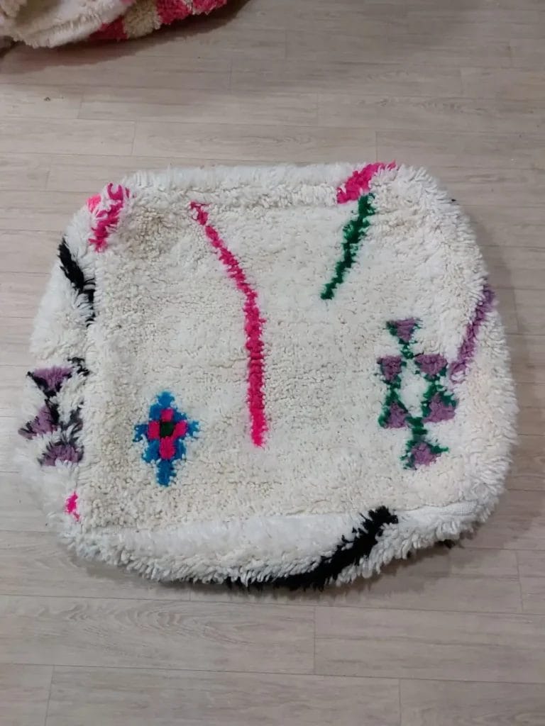 Een vierkante, donzige witte Marokkaanse Beni Ouarain Berber Poef 010 met kleurrijke abstracte patronen, waaronder vormen en lijnen in roze, blauw, groen, zwart en paars, liggend op een lichte houten vloer.