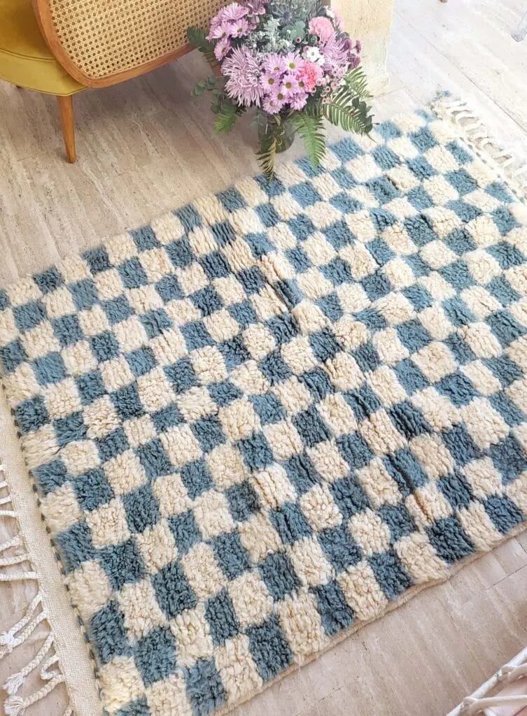 Een geblokt blauw en beige kleed op een lichte houten vloer, met een boeket roze en witte bloemen in een vaas ernaast.