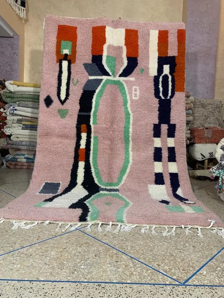 Een levendig, abstract patroon van 150 - 250 CM BENI OUARAIN VLOERKLEED 2166 MAROKKAANS TAPIJT HANDGEMAAKT met franjeranden rechtop weergegeven tegen een stapel gevouwen textiel op een tegelvloer.