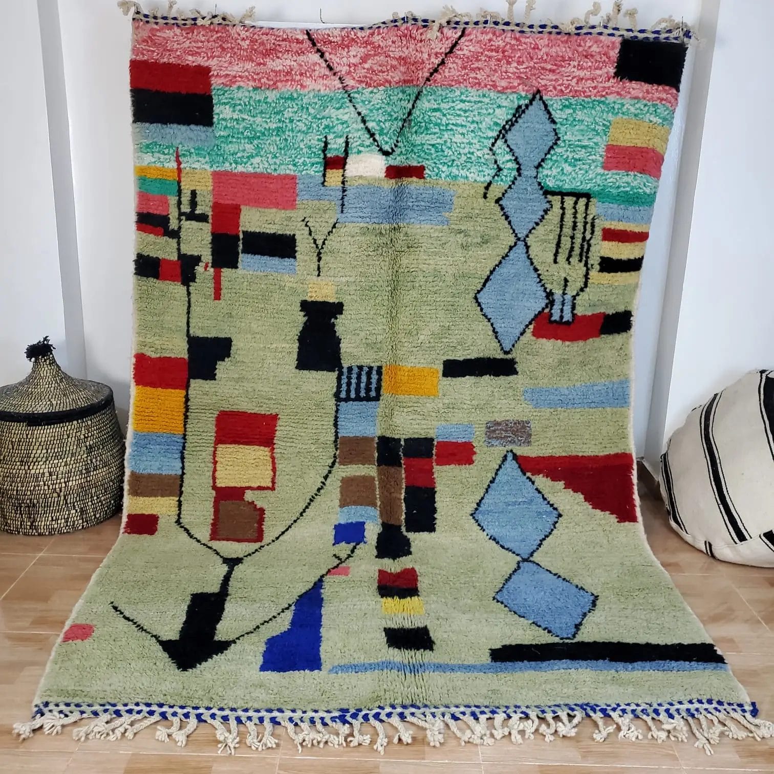 Een 100% HANDGEMAAKTE MAROKKAANSE TAPIJTEN TAPIJT 150 - 250 cm nr 113, versierd met abstracte geometrische patronen in rood, groen, blauw en zwart, staat tegen een muur met twee gevlochten manden aan de zijkanten.