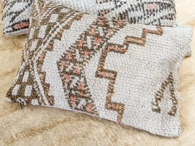 Een close-up van de Vintage Beni kussens - gebroken wit met textuur, zachtroze op een beige oppervlak heeft een ingeweven geometrisch patroon in bruin, groen en roze, dat perfect aansluit bij het aangrenzende vloerkleed.