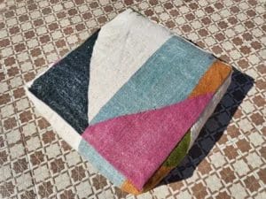 Een gevouwen, veelkleurige quilt met geometrische patronen ligt op een tegelvloer in het zonlicht en past prachtig bij het complexe ontwerp van de Marokkaanse Kilim Poef.