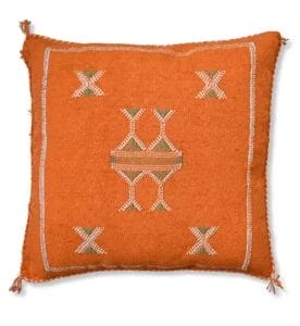 Het SABRA kussen van cactuszijde, kussen van Uksintas is een vierkant, oranje kussen versierd met symmetrische geometrische borduurpatronen in wit en groen. Met kwastjes op elke hoek. Het is een perfecte aanvulling op elk vloerkleed of tapijt in uw woonkamer.