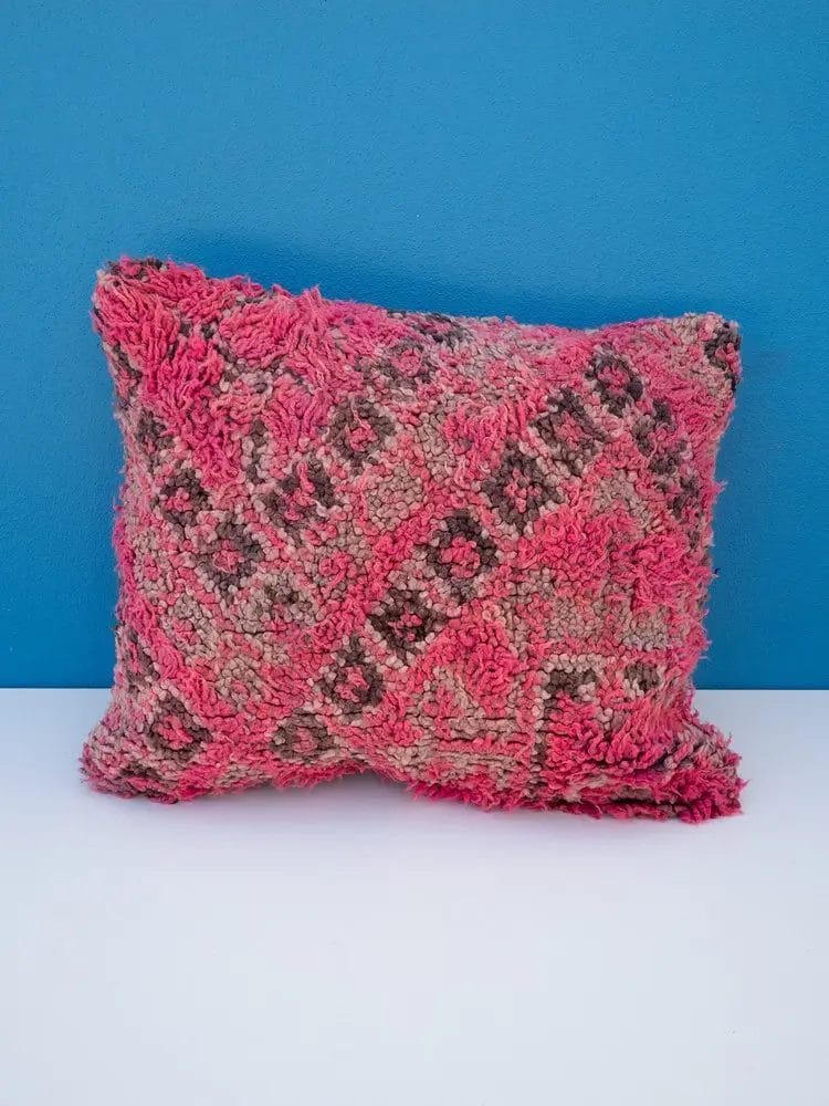 Een vierkant kussen, Handgemaakt kussen van Marokkaanse wol - 9 - 50 x 43 cm, met een gestructureerd, ruig oppervlak in tinten roze en grijs, rust tegen een blauwe muur op een wit oppervlak en complementeert de zachte tinten van het nabijgelegen tapijt.