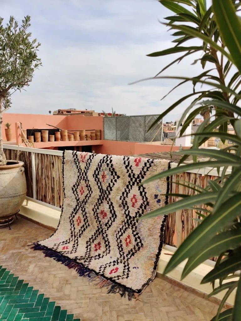 Een Ourika vloerkleed - rood detail met geometrische patronen is gedrapeerd over een bamboe balustrade op een zonnig terras. Er staan potplanten op de balustrade en een grote plantenbak aan de linkerkant. Een groene plant omlijst de rechterkant.