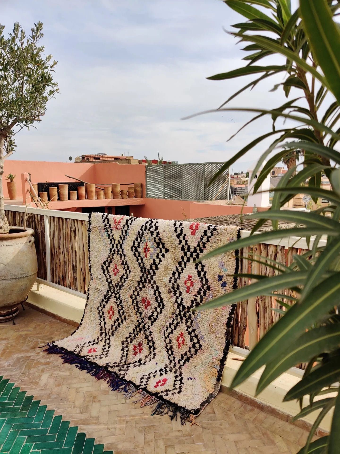 Een Ourika vloerkleed - rood detail met geometrische patronen is gedrapeerd over een bamboe balustrade op een zonnig terras. Er staan potplanten op de balustrade en een grote plantenbak aan de linkerkant. Een groene plant omlijst de rechterkant.