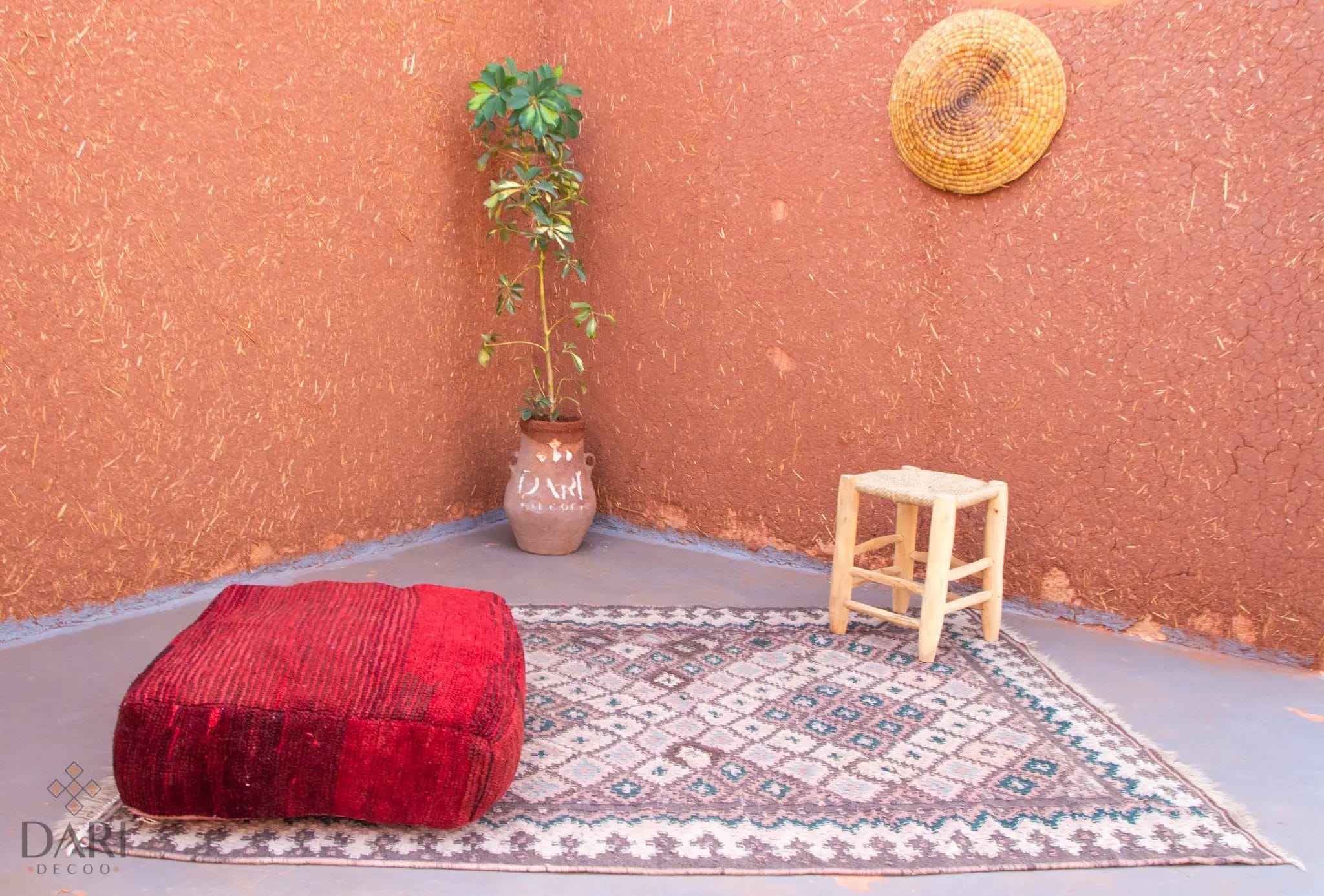 Een eenvoudig terrastafereel met een rood kussen, een potplant, een decoratief kleed, een klein krukje en een strohoed op een terracotta muur met structuur.