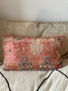 Een rechthoekig, geweven "Vintage kussens, Beni, roze en perzikkleur" kussen met een geometrisch patroon in roze, grijs en beige ligt op een neutrale bank en vormt een perfecte aanvulling op het aangrenzende vloerkleed.