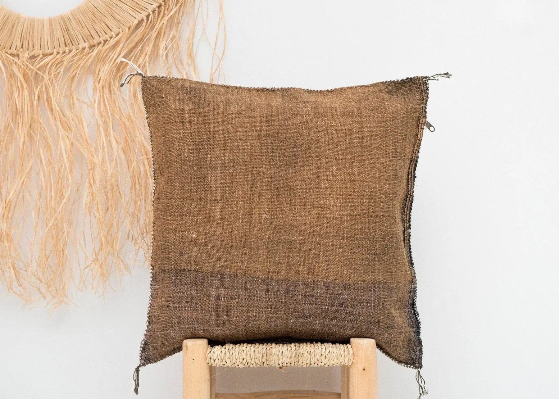 Een bruin jute kussen ligt op een houten stoel met franjeversiering aan een witte muur.