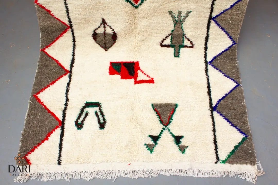 Een tapijt met geometrische en abstracte ontwerpen in rood, groen, blauw en zwart op een witte achtergrond, met zigzagranden en franje.