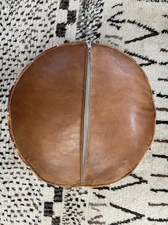 Een ronde, bruine leren poef met een rits in het midden, op een zwart-wit kleed met patroon.