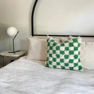 Een netjes opgemaakt bed met een Geruit Berberkussen, een handgemaakt wollen kussen met een geblokt groen en wit patroon, naast een klein rond nachtkastje met een lamp met een bolvormige witte lampenkap, ligt op een zacht tapijt dat de gezellige sfeer accentueert.