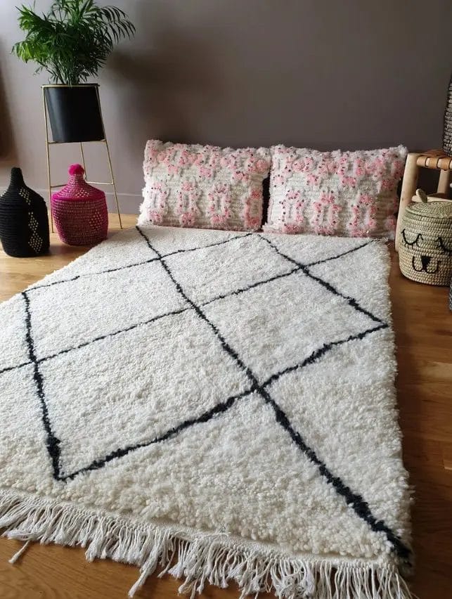 Een gezellige zithoek met twee bloemenkussens tegen de muur op een houten vloer, een wit hoogpolig vloerkleed met zwarte geometrische lijnen en een hoge plant in een zwarte pot ernaast.
