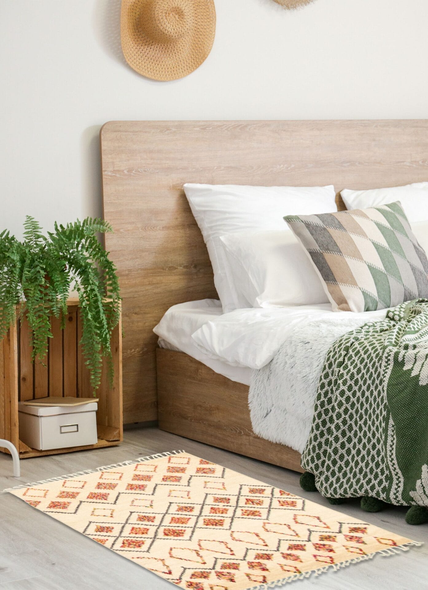 Een gezellige slaapkamer met een houten bed met wit en groen beddengoed, een geruit kussen, een varen in een houten plantenbak en een geometrisch kleed. Een strohoed hangt aan de muur.