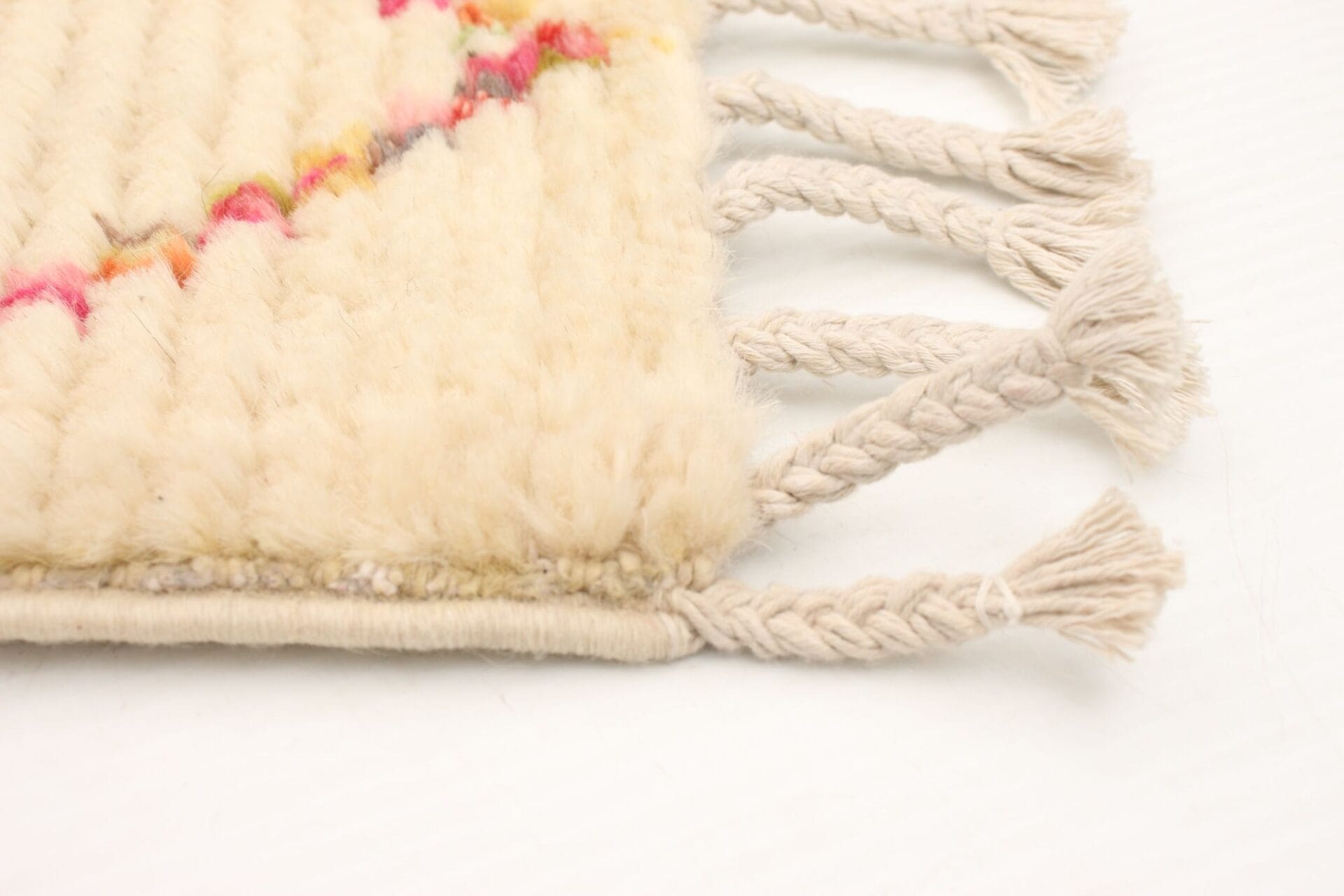 Close-up van een beige geweven hoekkleed met gevlochten franje en kwastjes, met een subtiel rood en roze diagonaal patroon.