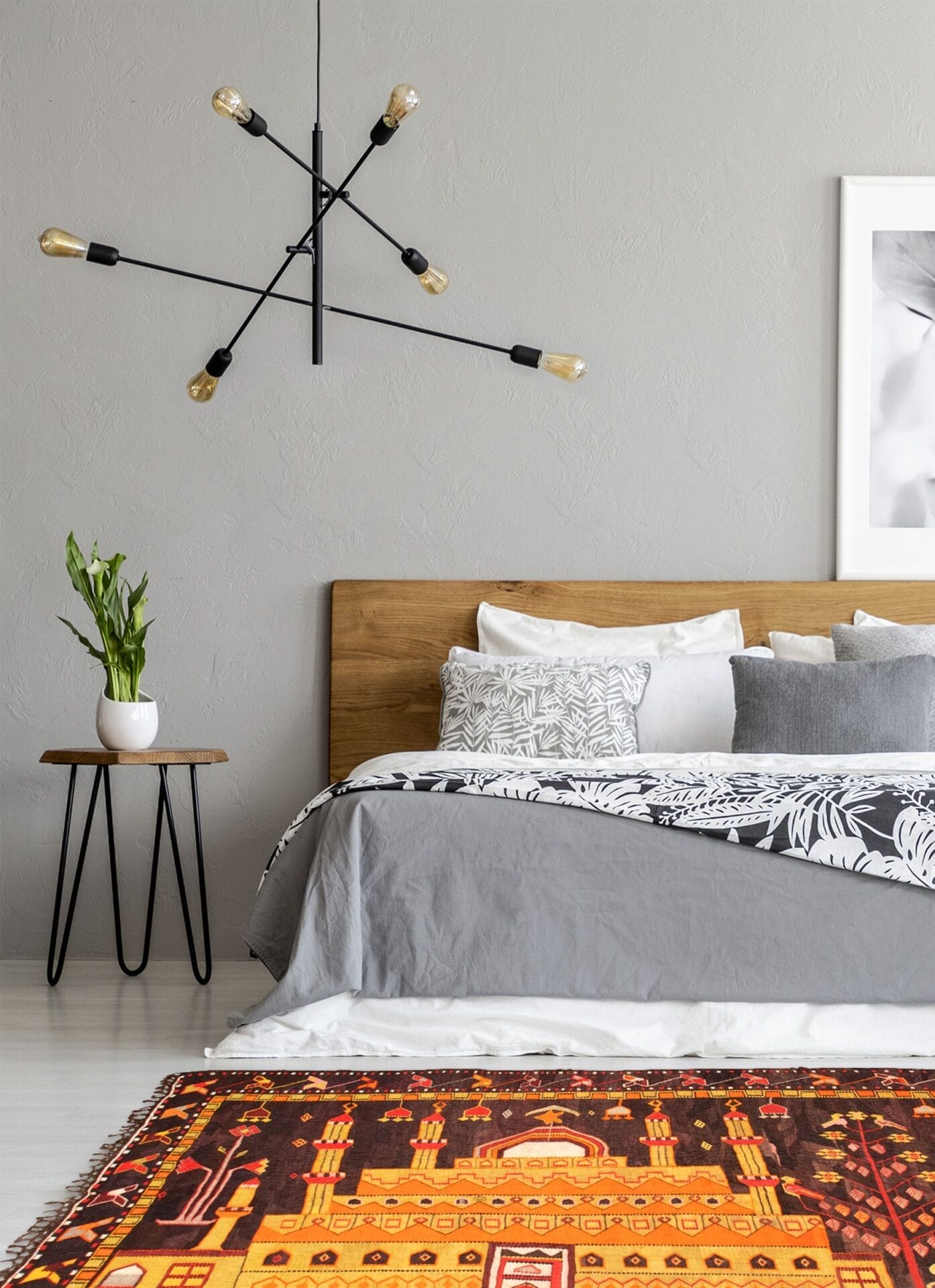 Een moderne slaapkamer met een houten bedframe, grijs en wit beddengoed, een vloerkleed met patroon en een bijzettafel met een potplant. Aan de muur hangen een eigentijdse plafondlamp en ingelijste kunstwerken.