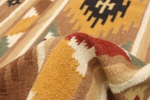 Close-up van een gestructureerd tapijt met geometrische patronen in tinten bruin, mosterdgeel, rood en zwart. Het tapijt is iets opgetild, waardoor het weefpatroon en de details zichtbaar zijn.