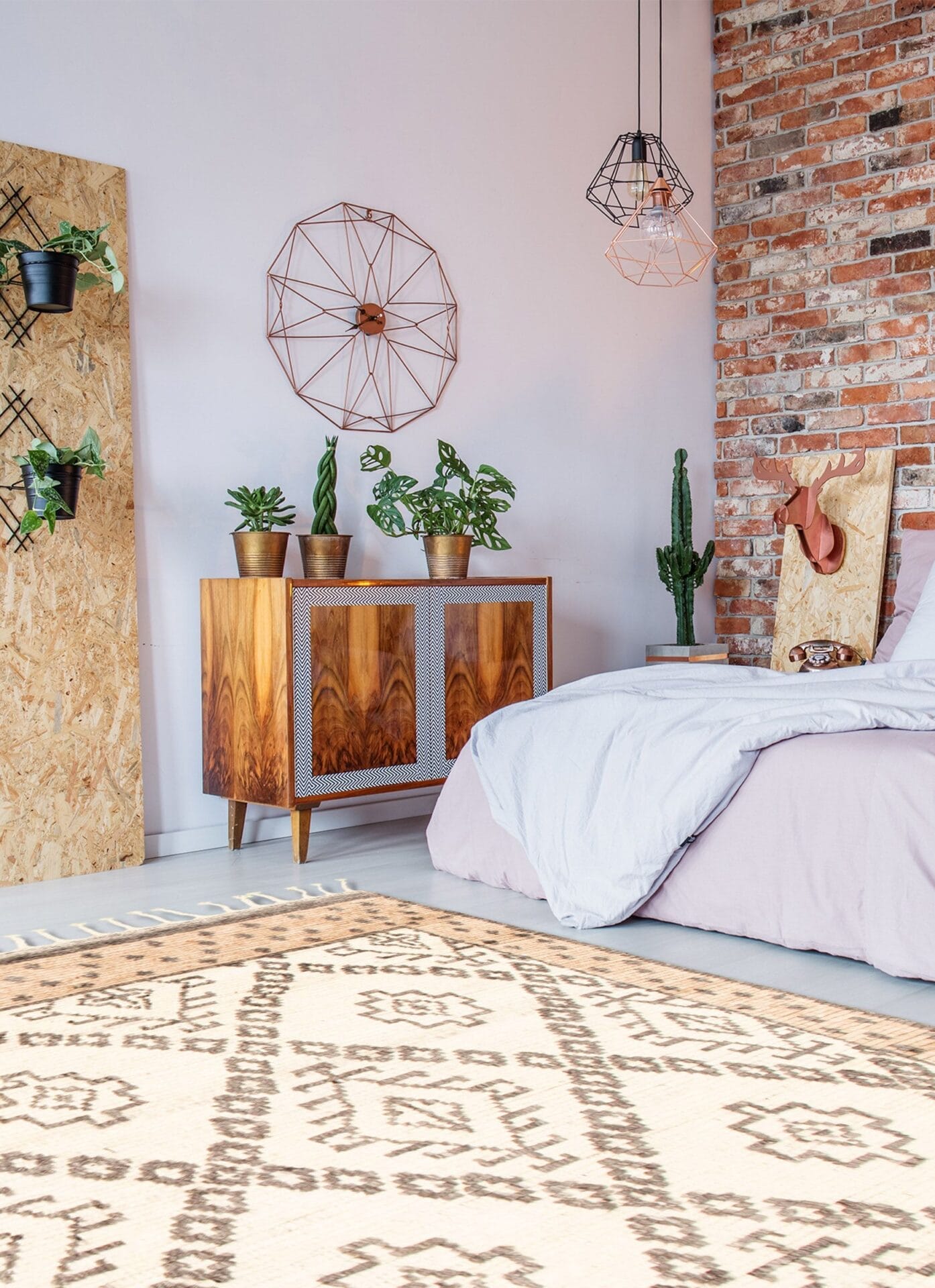 Een slaapkamer is voorzien van een bed met een lichtpaars dekbed, een houten kast met planten erop, geometrische wanddecoratie, hangende hanglampen, een bakstenen muur en een beige vloerkleed met donkerbruine patronen.