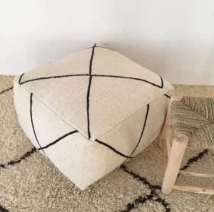 Een beige, vierkante Marokkaanse Berber-poef met zwarte kruisende lijnen staat op een gestructureerd tapijt naast een houten kruk met een geweven zitting. Het geheel staat elegant tentoongesteld op een zacht tapijt.