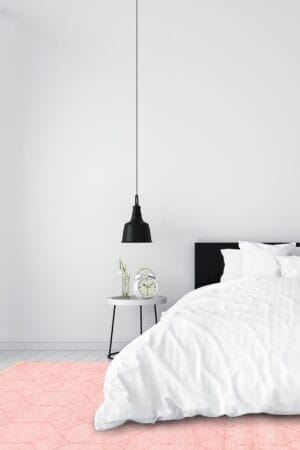 Minimalistische slaapkamer met witte muren, een wit bed en een zwart hoofdeinde. Op een klein rond nachtkastje staan een witte wekker en een plant. Boven de tafel hangt een zwarte hanglamp en op de vloer ligt een roze vloerkleed.