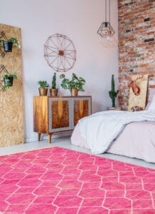 Een moderne slaapkamer met een bakstenen muur is voorzien van een bed, een houten kast, aan de muur gemonteerde planten en een geometrische inrichting. Een felroze vloerkleed met een wit patroon bedekt de vloer.