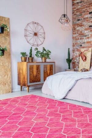 Een moderne slaapkamer met een bakstenen muur is voorzien van een bed, een houten kast, aan de muur gemonteerde planten en een geometrische inrichting. Een felroze vloerkleed met een wit patroon bedekt de vloer.