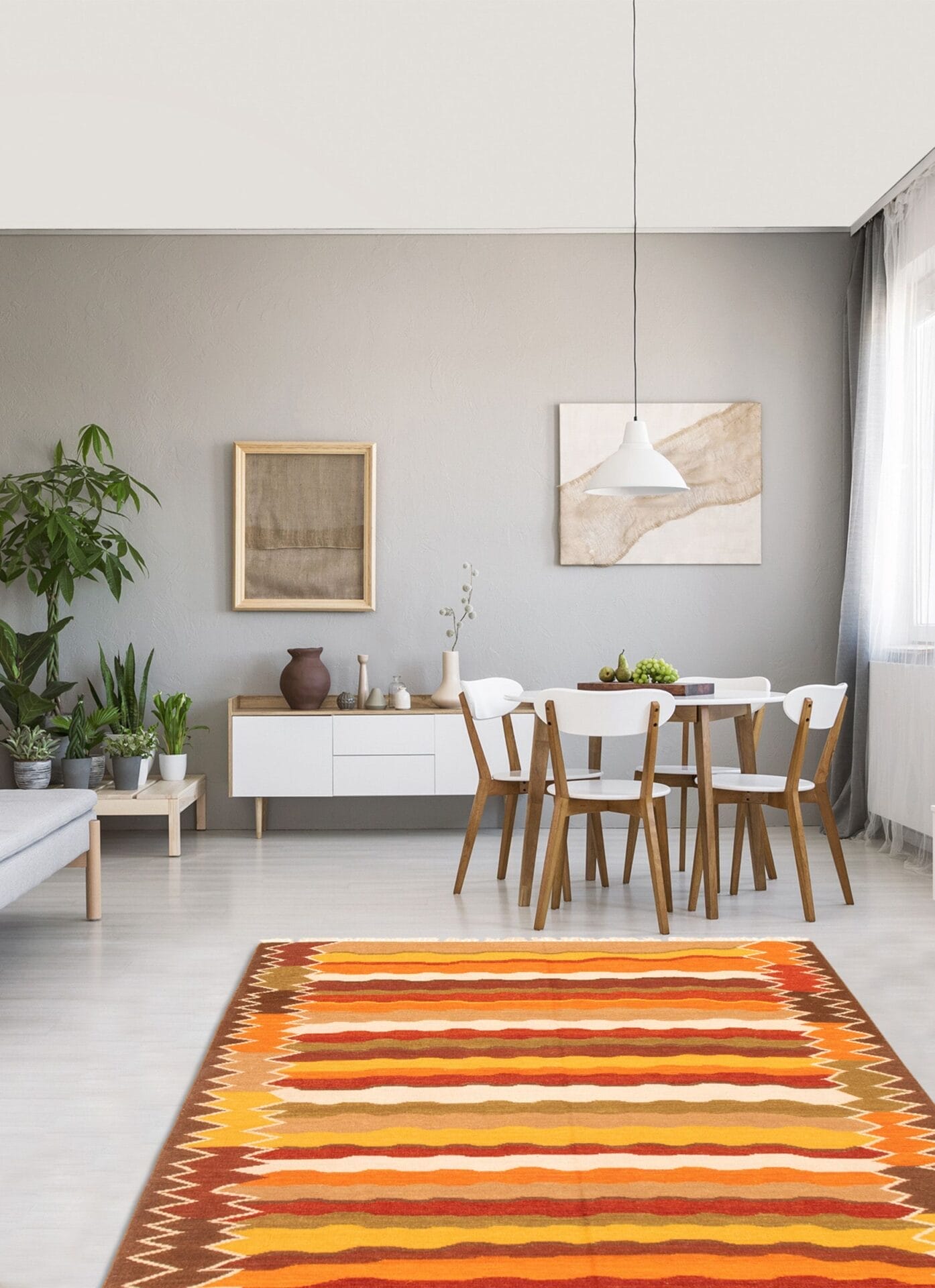 Een moderne eetkamer met een houten tafel en stoelen, een wit dressoir, potplanten, kunstwerken aan de muren en een veelkleurig vloerkleed op een lichte vloer. Boven de tafel hangt een witte hanglamp.