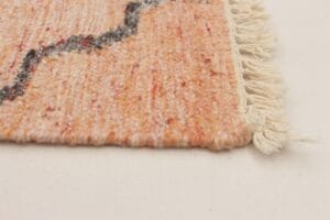 Close-up van een beige kleed met rode accenten, met een zwarte golvende lijn. De franje is zichtbaar aan de rand.