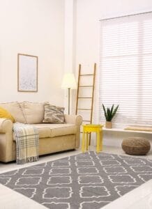 Woonkamer met een beige bank, een kussen met patroon en een geel bijzettafeltje. Een lamp en een ladder staan tegen een muur met jaloezieën. Een plant en een poef staan op de lichte houten vloer naast een geometrisch kleed.