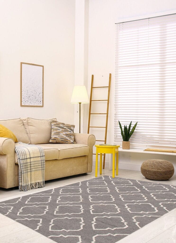 Woonkamer met een beige bank, een kussen met patroon en een geel bijzettafeltje. Een lamp en een ladder staan tegen een muur met jaloezieën. Een plant en een poef staan op de lichte houten vloer naast een geometrisch kleed.