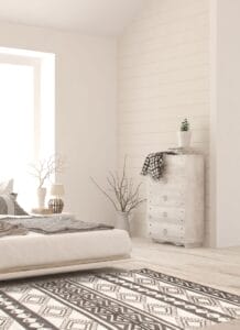 Een minimalistische slaapkamer met een laag bed, een ladekast met een plant erop, een vloerkleed met geometrisch patroon en lichtinval door een groot raam. Een deken en takken in een vaas maken deel uit van het decor.