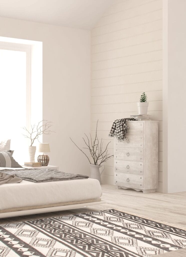 Een minimalistische slaapkamer met een laag bed, een ladekast met een plant erop, een vloerkleed met geometrisch patroon en lichtinval door een groot raam. Een deken en takken in een vaas maken deel uit van het decor.