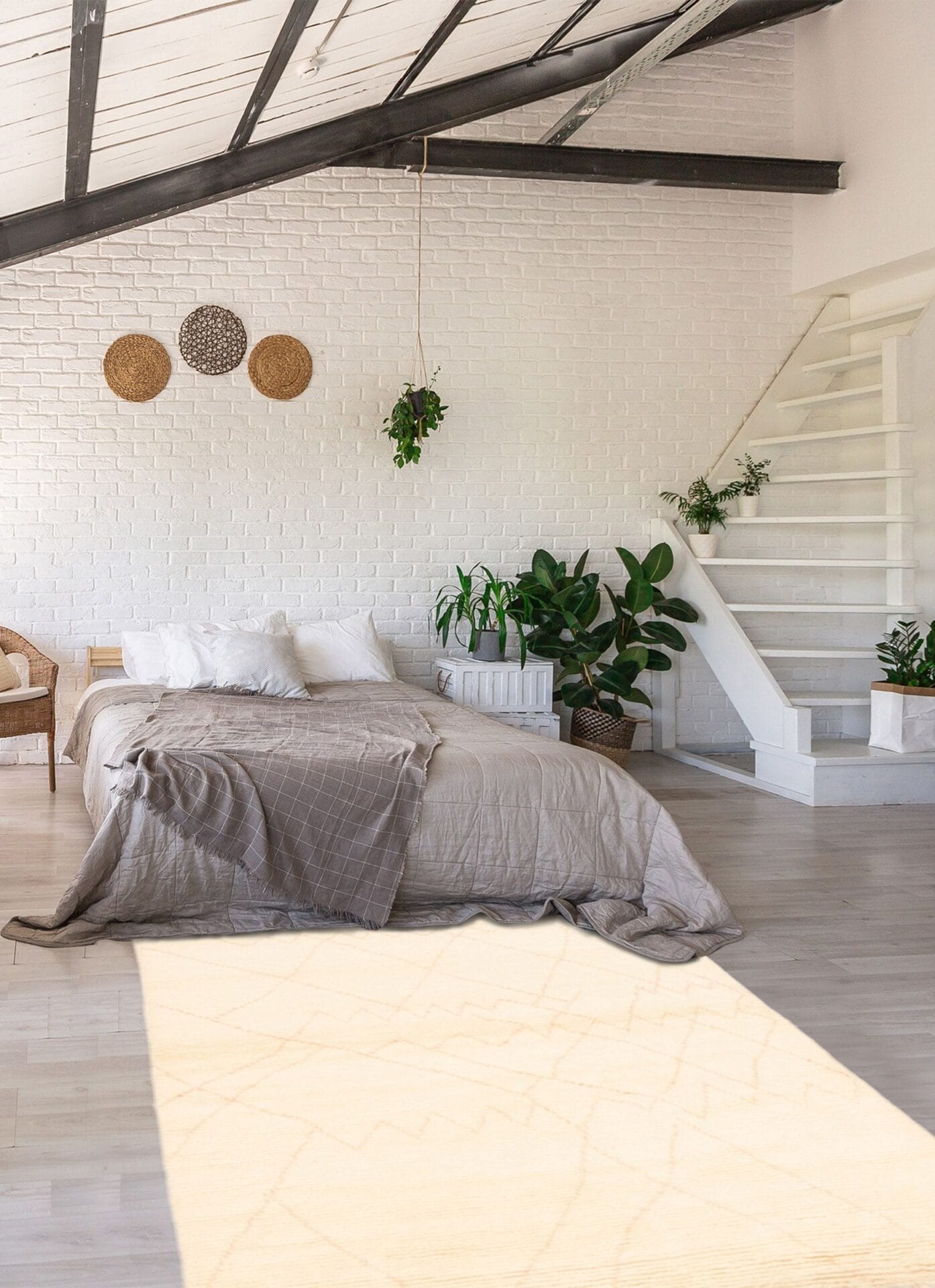 Zonovergoten slaapkamer met een schuin plafond, witte bakstenen muur en bed met beige beddengoed. Inclusief planten, rieten stoel en hangende decoraties. Witte trap die naar boven leidt. Lichte houten vloer geaccentueerd door een zacht tapijt.