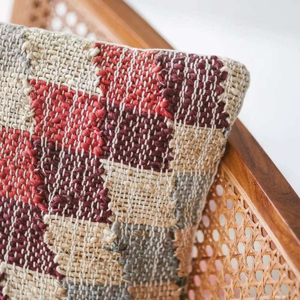 Een geweven kussen met een rood, bruin en beige schaakbordpatroon rust op een houten stoel met een rotan rugleuning.