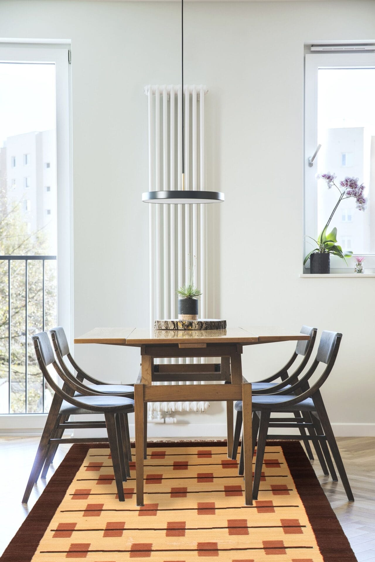 Moderne eethoek met een houten tafel, zwarte stoelen, een vloerkleed met geometrisch patroon en een hangende lamp. Op tafel staat een potplant, met extra planten bij het raam op de achtergrond.