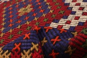 Close-up van kleurrijk geweven textiel, dat lijkt op een traditioneel tapijt, met een geometrisch patroon met rode, blauwe, groene, witte en gele draden gerangschikt in een rasterachtig ontwerp.