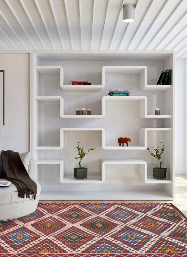 Tegen een muur staat een moderne witte zigzagboekenplank met diverse boeken, potplanten en decoratieartikelen. Op de voorgrond staat een vloerkleed met patronen en een gedeeltelijk zichtbare lichtgrijze stoel.