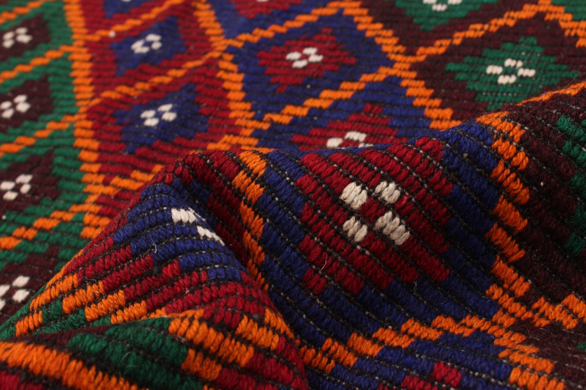 Close-up van kleurrijk geweven textiel dat lijkt op een tapijt of vloerkleed, met geometrische patronen in rood, blauw, groen, oranje en geel met witte accenten. De stof heeft een licht verhoogde, gestructureerde uitstraling.