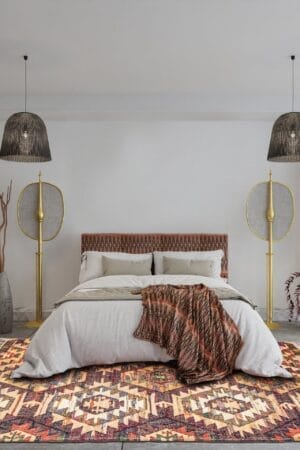 Een moderne slaapkamer met een groot bed, hoofdeinde met patroon, wit beddengoed, een kleurrijk vloerkleed en unieke gouden vloerlampen. Aan het plafond hangen twee hanglampen.