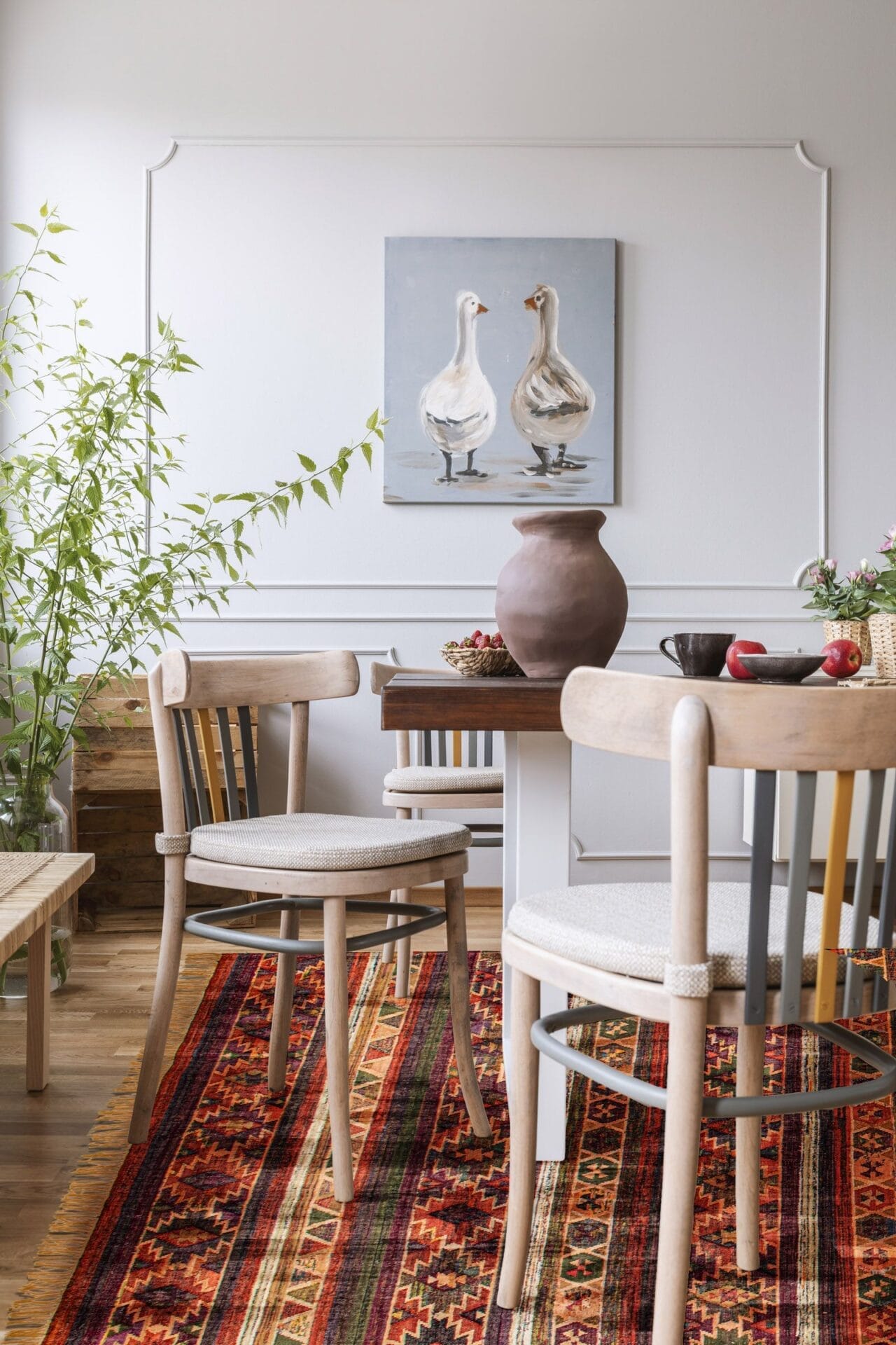 Eethoek met een houten tafel, twee stoelen, een kleivaas en een kleurrijk vloerkleed. Aan de muur hangt een schilderij van twee witte eenden. In de hoek wordt een plant geplaatst.