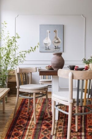 Een eethoek met twee stoelen, een ronde tafel, een vaas en serviesgoed. Aan de muur hangt een schilderij van twee ganzen. Aan de linkerkant staat een potplant en op de vloer ligt een tapijt met kleurrijke patronen.