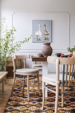 Een eethoek met een houten tafel, twee stoelen, een kleurrijk vloerkleed, planten en een schilderij van twee eenden aan de muur.