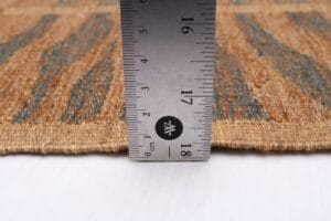 Een metalen liniaal meet de dikte van een vloerkleed van geweven stof. De liniaal geeft aan dat de dikte van het vloerkleed ongeveer 18 millimeter of 1,8 centimeter is. Het vloerkleed heeft een structuurpatroon.