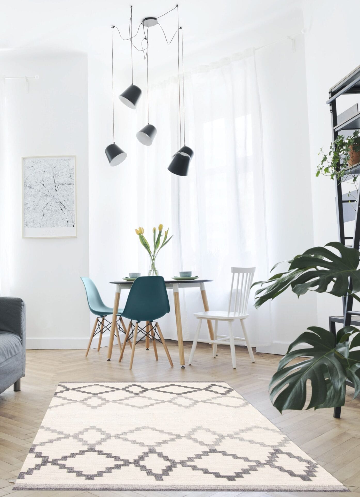 Een moderne, lichte kamer met een kleine eettafel, vier stoelen, een vloerkleed met geometrisch patroon, een potplant en een zwarte hanglamp. Witte gordijnen laten natuurlijk licht de ruimte vullen.